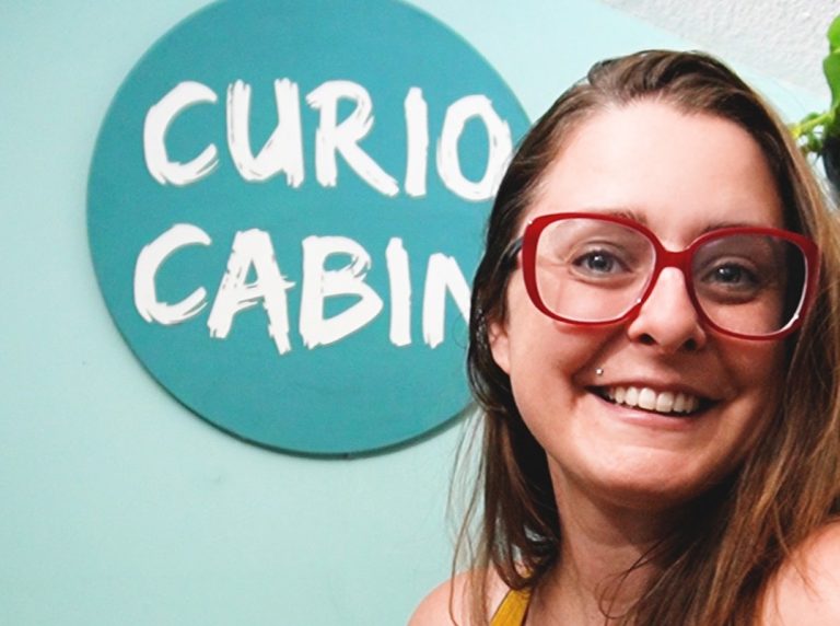 a woman with light skin and medium brown hair is smiling holding hand hand out.. She has red glasses and a mustard yellow tank top. She's standing infront of a laser cut wood sign hanging on a wall that says Curio Cabin in white script font on a teal background. The wall is a light tone of teal.