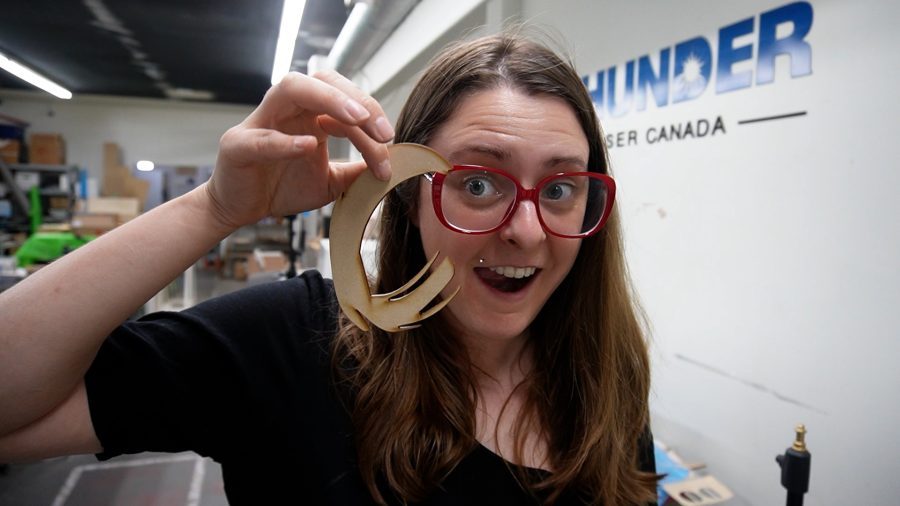 a young light skinned woman with long light brown hair holds a laser cut letter made of wood. In the background the words Thunder Laser Canada are on the wall