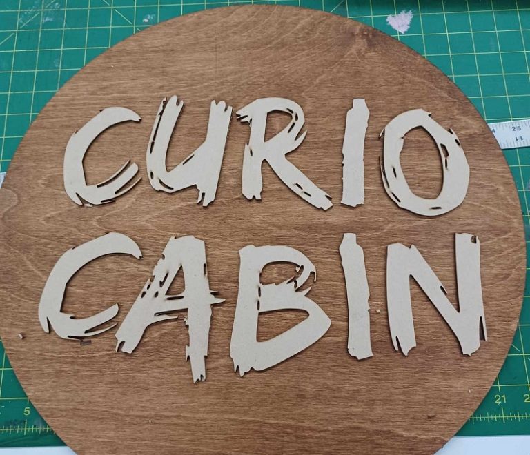 circular wood sign with laser cut letter made from mdf. Letters are a brush script and say Curio Cabin. They are made on a Thunder Laser machine
