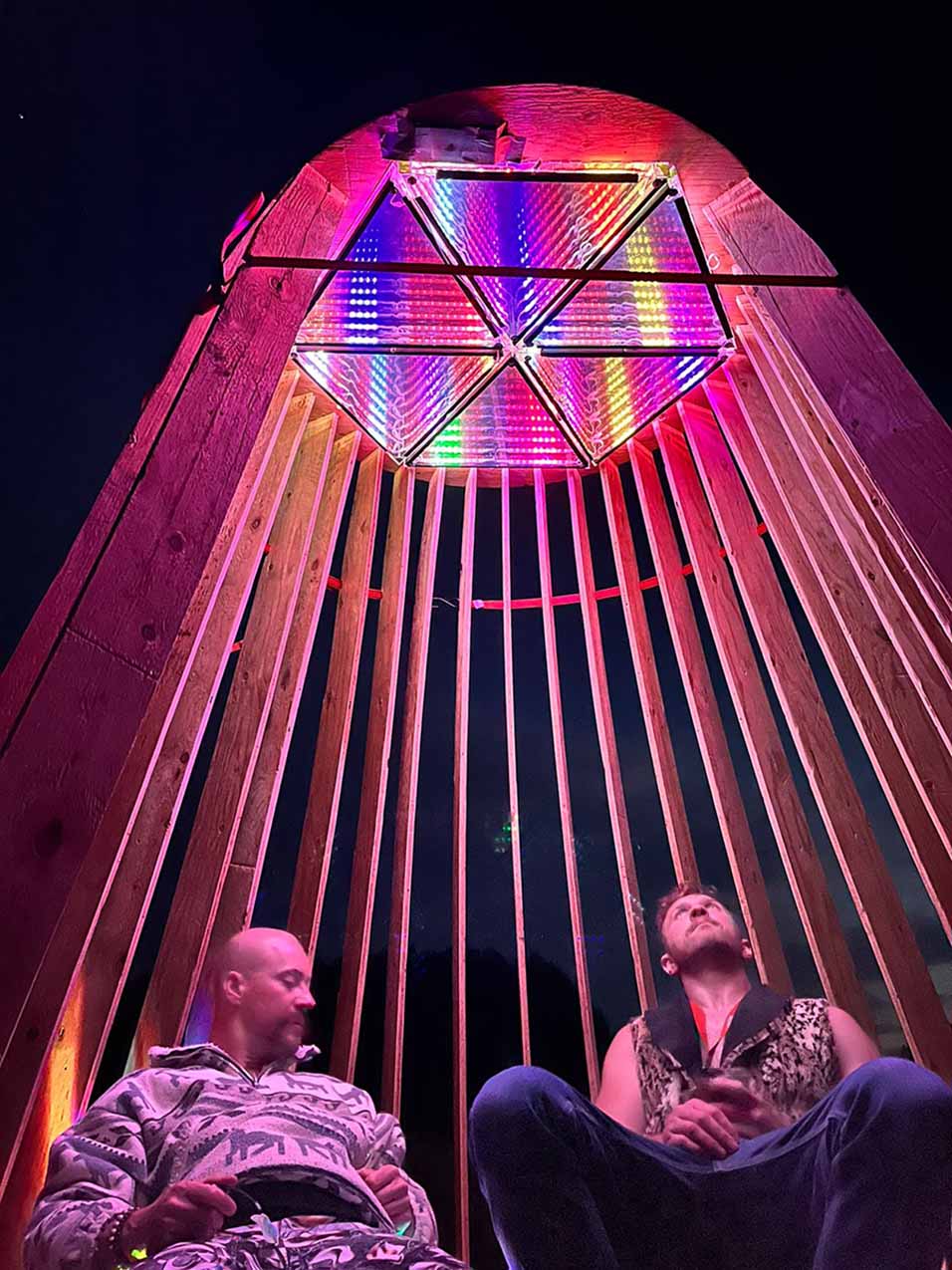 2 people sit under a cone shaped wood structure with an inifinty mirror ontop. One guy is looking up at it. There are pink and purple and white lights in the mirror illuminating them