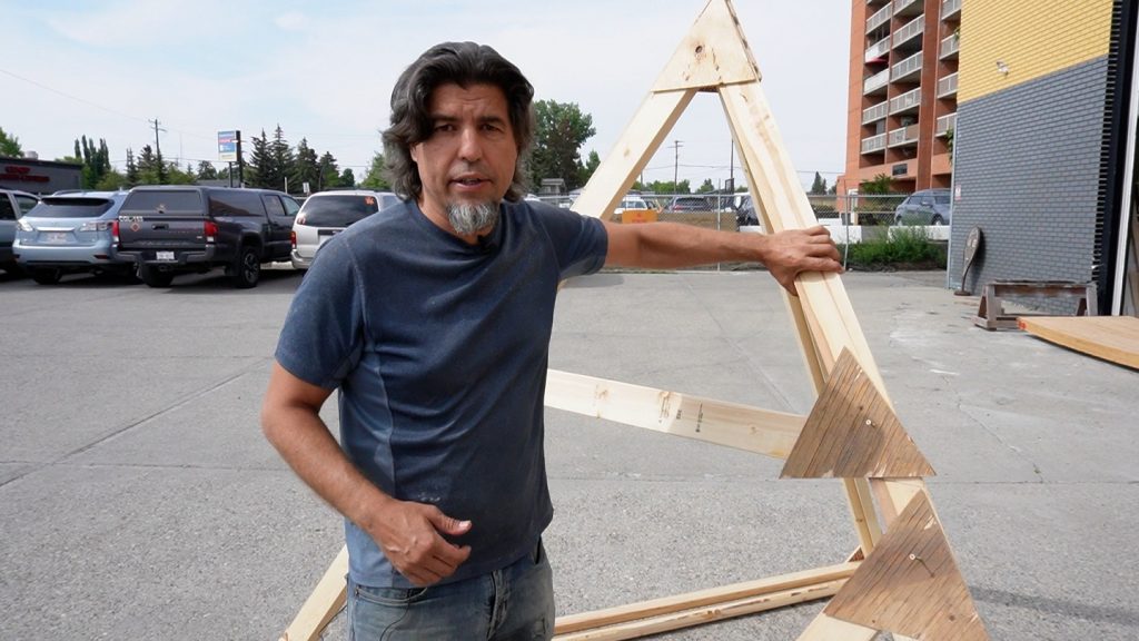 Artist Ken Hacke stands outside in a parking lot with a wooden triangle structure. Ken is a middle aged man with a medium to dark skin tone with ear length dark hair and a short grey goatee.
