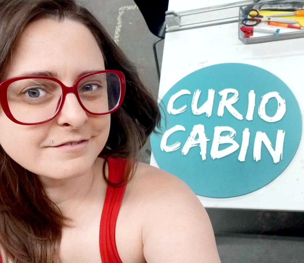 a light skinned woman with dark hair and red glasses smiled taking a selfie of herself and her laser cut wood sign. behind her is the sign she made on a laser cutter that says Curio Cabin-a round teal sign with white brush script font.