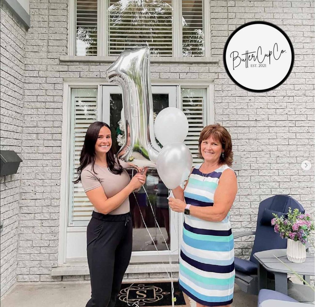 2 women stand infront of a house holding a silver balloon shaped like a number 1. Left is Danielle smith, daughter, righ tis Susan Smith, mother both owners of Buttercup Co Laser Engraved drinkware
