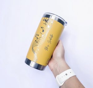 A hand holding a yellow metal laser engraved coffee mug tumbler. Engraved on it is a music staff and notes and the name of a music teacher. white background