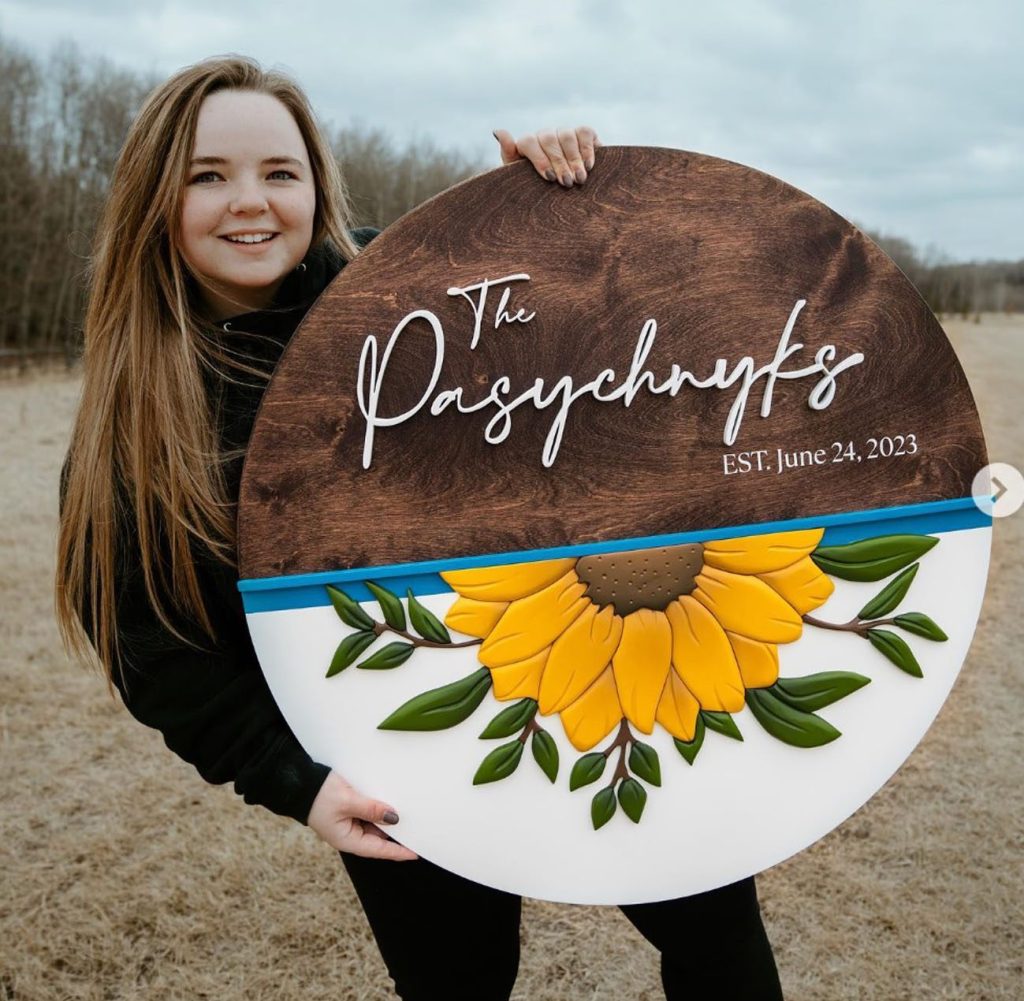 A light skinned girl with brownish blond hair holds a large sign. Half is wood grain and half has a large yellow sign flower. the edges of the sunflower. TThe top half says "The Pasychnyks"