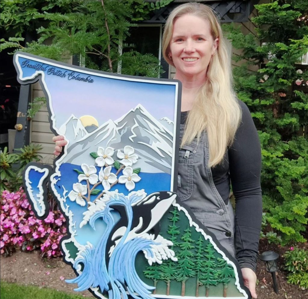 A kight skinned woman with long blond hair holds a sign in the shape of the province of BC.