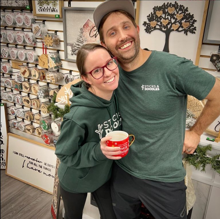 a light skinned man (Kyle)and woman (Danelle) in their 30's smile standing inf front of a display rack of wood laser cut ornaments. They are wearing green shirts with the logo Sticks and Doodles small on the front.