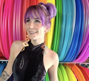a light skinned woman with light purple hair smiles standing in front of rainbow colored streamers