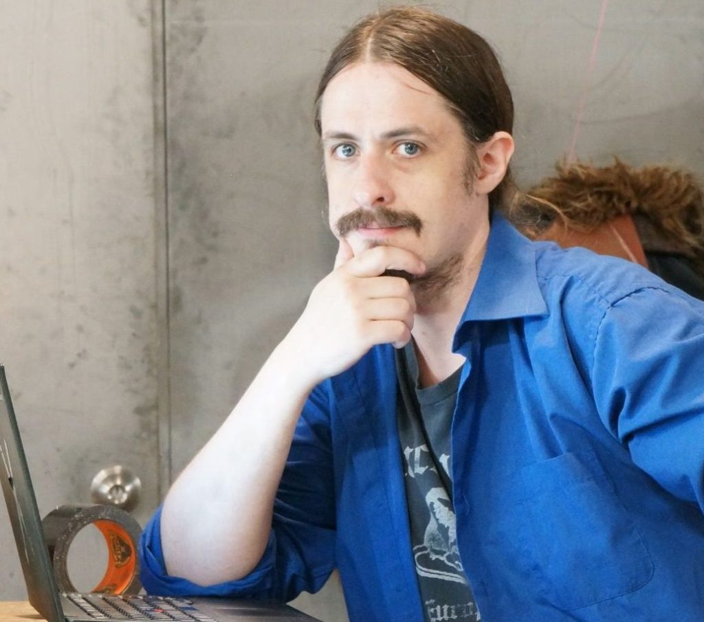 a light skinned man white a ponytail sits at a desk with his arm on the table and hand on his chin with a laptop infront of him