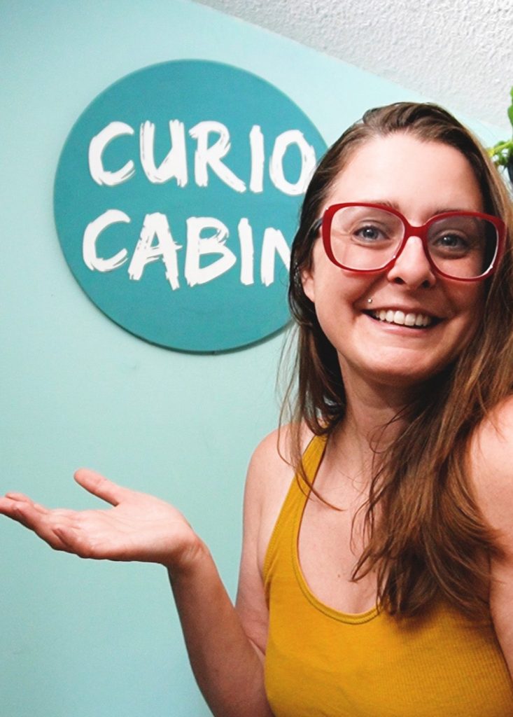 ee stands in front of a sign that says Curio Cabin. Shes got long brown hair and wearing large red glasses and a yellow shirt