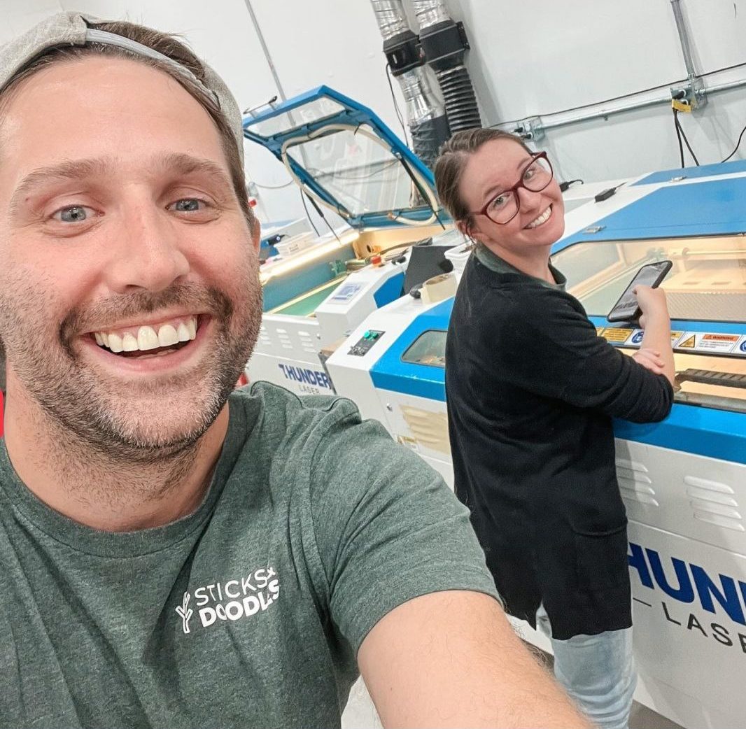 a light skinned man is wearing a backwards baseball hat smiling an holding the camera. He is wearing a branded green shirt that says Sticks and Doodles in white. In the background a woman is smiling and standing in front of a Thunder Laser laser cutter machine. She is light skinned wearing her hair in a bun and has thick rimmed glasses.