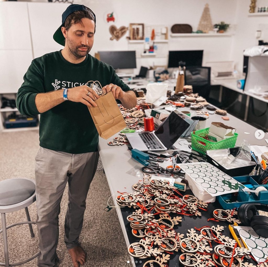 kyle- a man in pants and a long sleeve shirt and a backwards hat standing infront of a table covered in christmas ornaments