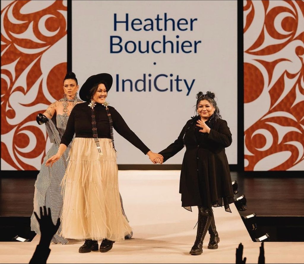 two women stand holding hands on a runway at Vancouver indigenous fashion week
