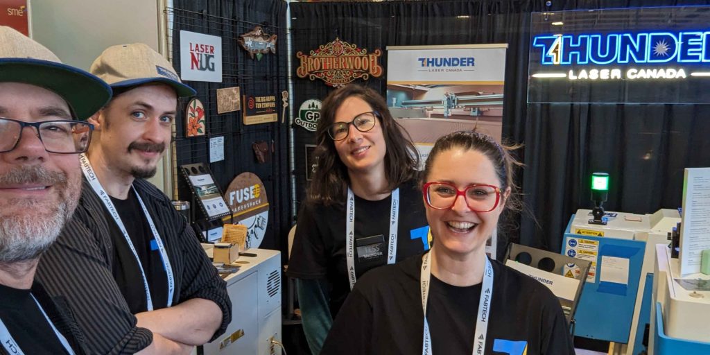 thunder laser canada team smiling at a conference event with wooden and acrylic signs signs made by customers in the background on display customers