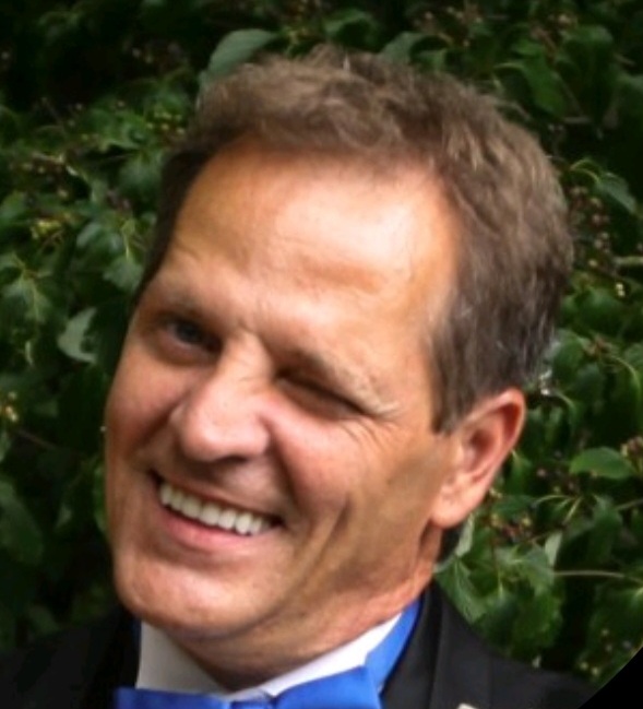 a light skinned man wearing a suit winking. He has short brown hair and is clean shaven